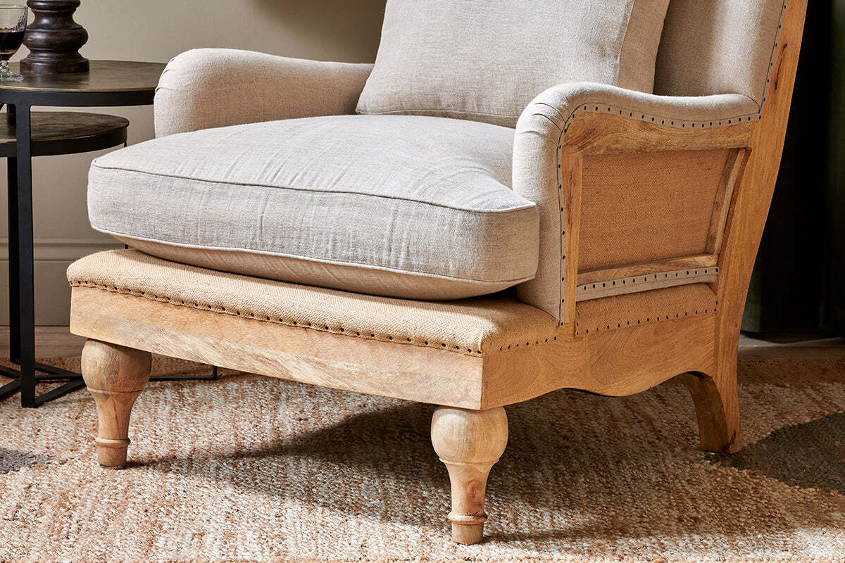 A close-up of the nkuku Abe Deconstructed Linen Armchair in Stone, featuring light beige linen upholstery, nailhead trim, a thick cushioned seat, and carved mango wood frame. The chair sits on a textured rug beside a small round table.