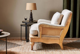 The Abe Deconstructed Linen Armchair - Stone by nkuku, featuring a beige linen upholstery and mango wood frame, sits on a patterned rug next to two round nesting tables with a dark wooden lamp, red wine glass, dark green curtain, and neutral wall in the background.
