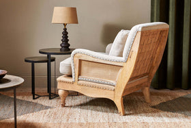 The nkuku Abe Deconstructed Linen Armchair in Stone rests on a woven rug next to two round black nesting tables and a brown lamp. The room features neutral walls and dark green curtains.