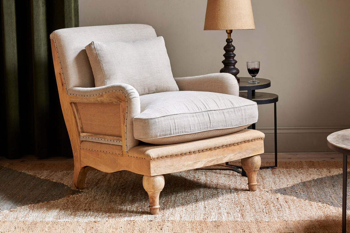 The nkuku Abe Deconstructed Linen Armchair in Stone sits on a woven rug with a matching cushion. Beside it, a small round side table holds a lamp and glass of red wine. A green curtain and light-colored wall form the backdrop.