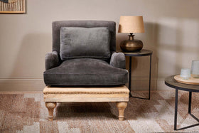 The nkuku Abe Deconstructed Velvet Armchair in moss, featuring a matching back cushion, pairs with a light wood ottoman and round side table topped by a textured brown lamp, all resting on a patterned beige rug against a neutral wall.