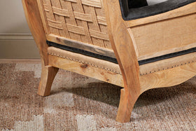 Close-up of the Abe Deconstructed Velvet Armchair in Moss by nkuku, featuring curved legs, velvet upholstery, and black nailhead trim. The chair rests on a beige and cream geometric rug against a light wall and baseboard.