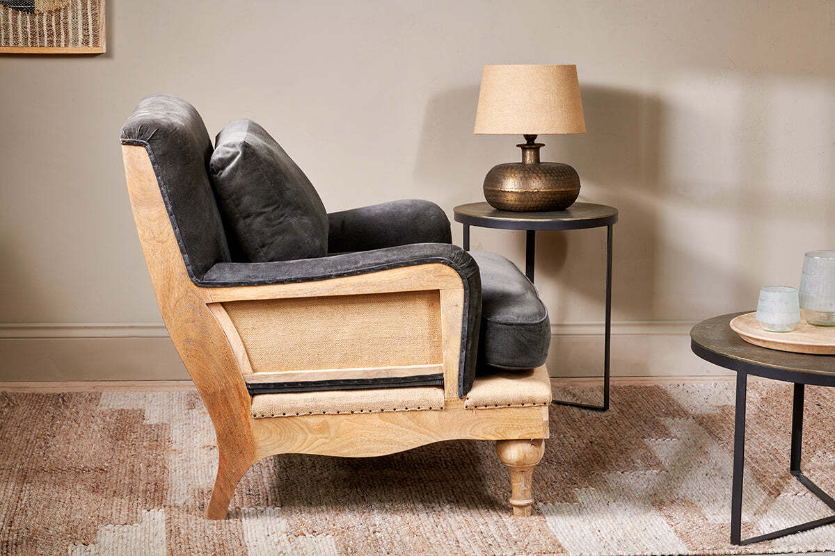 The Abe Deconstructed Velvet Armchair in moss by nkuku sits on a patterned rug. Next to it, a round black side table holds a bronze lamp with a beige shade, while another table displays glassware. The wall behind is soft beige.