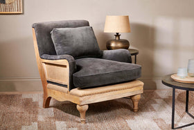 The Abe Deconstructed Velvet Armchair by nkuku in moss sits on a patterned rug with dark gray cushions. A round side table with a lamp and glassware stands nearby, while a light beige wall enhances the cozy, inviting atmosphere.