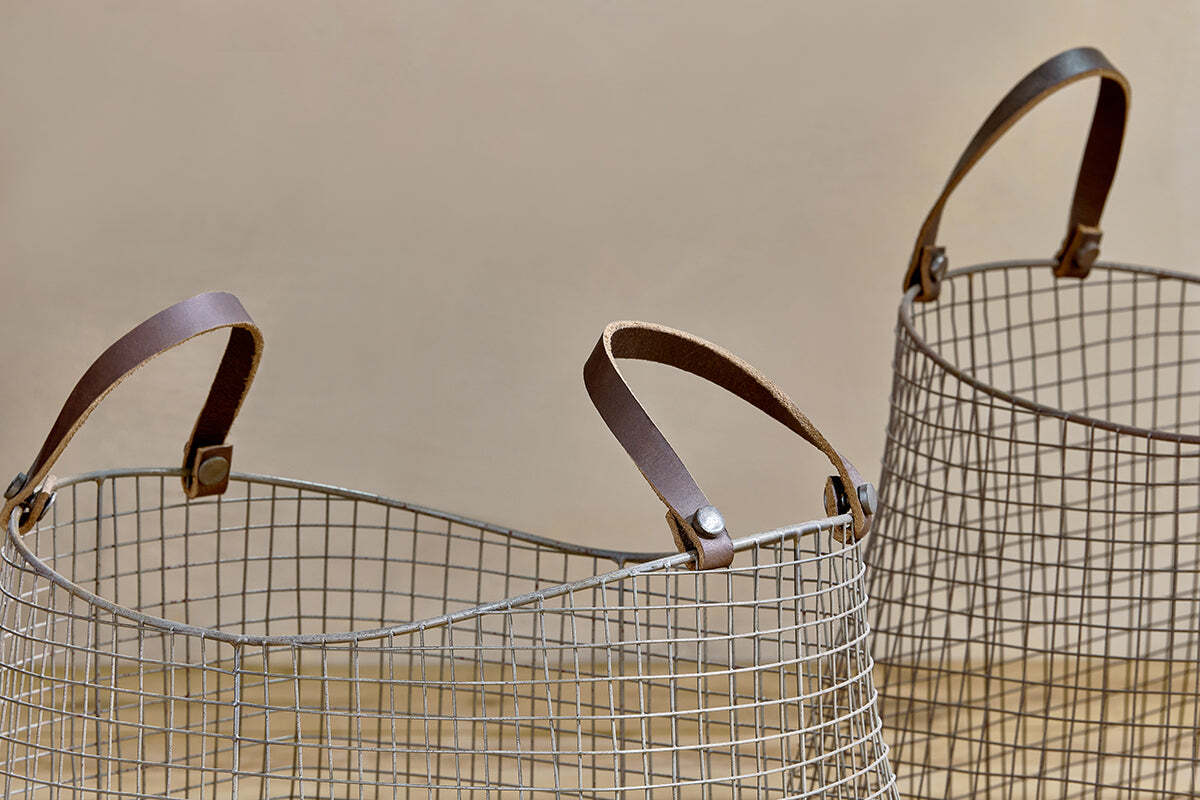 The Adah Iron Baskets - Brown (Set of 2) by nkuku sit on a light wooden surface, showcasing their open grid design and sturdy brown leather handles—perfect stackable storage with only the tops and handles visible.