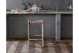 The Adembi Woven Leather Counter Stool by nkuku, with its wooden frame, stands on a herringbone floor near a gray concrete kitchen counter topped with dishes, glassware, potted plants, accessories, and a modern faucet in the background.