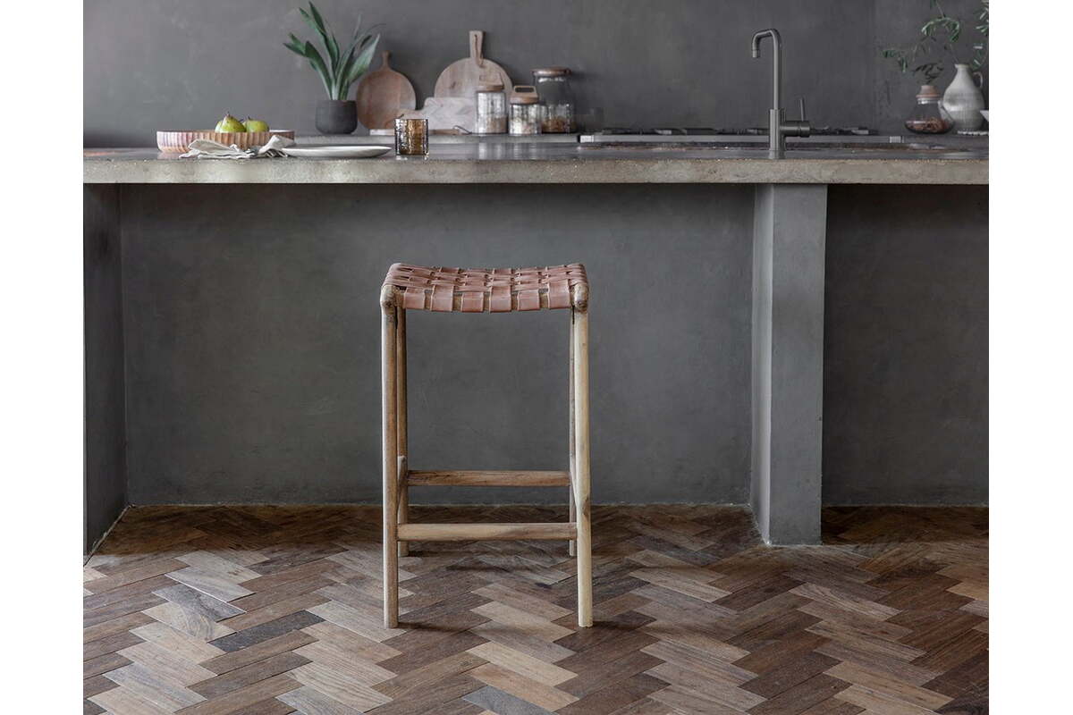 The Adembi Woven Leather Counter Stool by nkuku, with its wooden frame, stands on a herringbone floor near a gray concrete kitchen counter topped with dishes, glassware, potted plants, accessories, and a modern faucet in the background.
