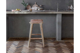 The Adembi Woven Leather Counter Stool by nkuku sits beside a concrete countertop in a modern kitchen with gray walls, chevron wood flooring, and minimalist decor featuring plants, fruit, and cutting boards.