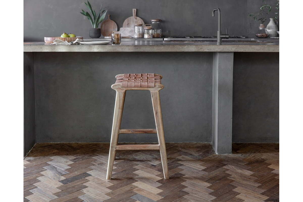The Adembi Woven Leather Counter Stool by nkuku sits beside a concrete countertop in a modern kitchen with gray walls, chevron wood flooring, and minimalist decor featuring plants, fruit, and cutting boards.