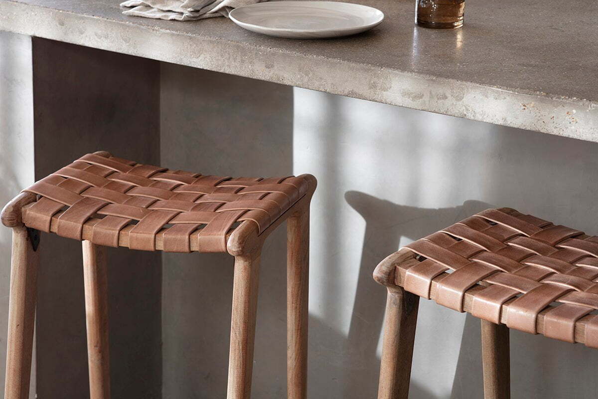 Two nkuku Adembi Woven Leather Counter Stools with tan seats are placed by a gray stone countertop, which holds a white plate, brown glass, and folded napkin, all illuminated by natural light that casts gentle shadows.