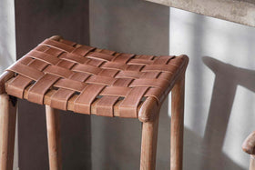 A close-up of the nkuku Adembi Woven Leather Counter Stool shows its tan, tightly woven leather seat in a checkered pattern atop rounded light acacia wood legs, with soft light casting gentle shadows on the neutral gray wall behind.