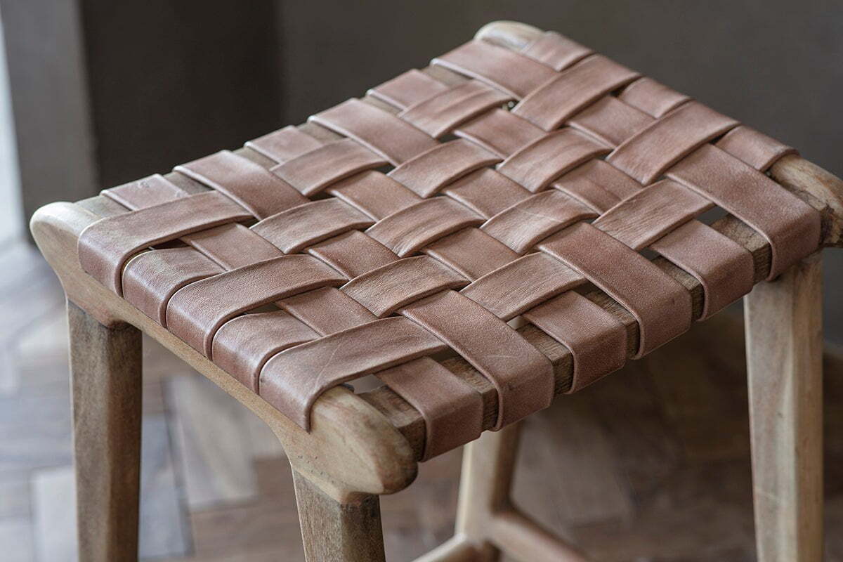 A close-up of the nkuku Adembi Woven Leather Counter Stool, showcasing wide tan leather straps in a crisscross pattern and a natural-finish wood frame, set on an elegant herringbone hardwood floor.