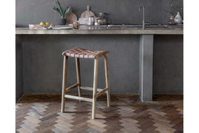 A rustic kitchen showcases nkuku's Adembi Woven Leather Counter Stool with a wooden frame. A concrete countertop, sink, kitchen items, and a plant in a vase complete the cozy space, while a herringbone wood floor adds warmth.