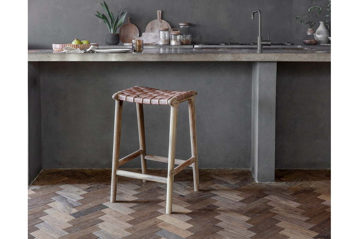 A rustic kitchen showcases nkuku's Adembi Woven Leather Counter Stool with a wooden frame. A concrete countertop, sink, kitchen items, and a plant in a vase complete the cozy space, while a herringbone wood floor adds warmth.