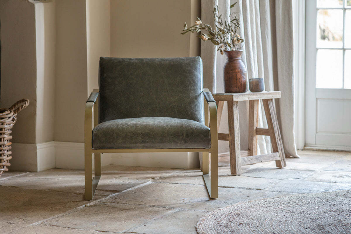 The nkuku Adra Leather & Brass Occasional Chair in moss green sits on a stone floor. To the right is a wooden side table with a vase of dried flowers and a small dark pot.