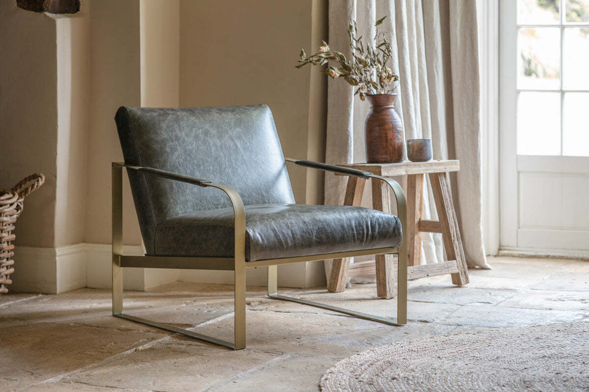 The nkuku Adra Leather & Brass Occasional Chair in moss green sits on a stone tile floor by a window with beige curtains. Next to it, a rustic wooden stool displays a brown vase of dried branches, filling the cozy living space with natural light.