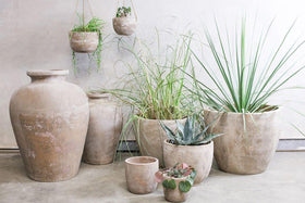 A group of rustic garden decor, including the nkuku Affiti Clay Planter - Medium, sits on a concrete floor against a light wall. Filled with grasses and succulents, two smaller handmade pots hang above the larger planters.