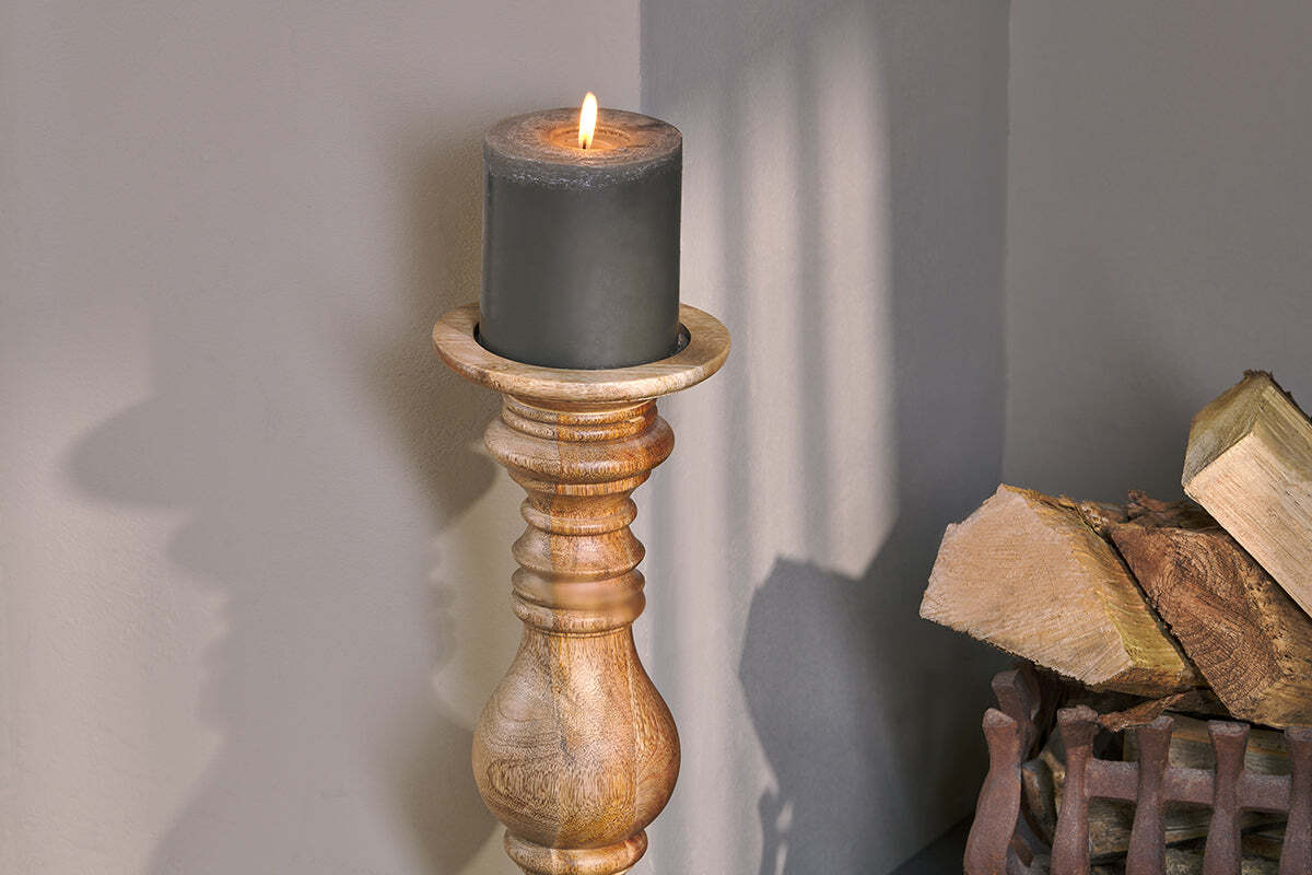 A lit black candle sits on the Akello Candle Stick by nkuku. Firewood in a metal holder rests nearby, while light and shadow dance on a gray wall, creating a cozy, rustic setting ideal for fireplace decor.
