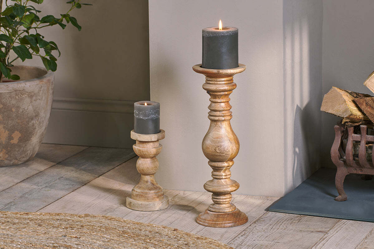 Two lit dark gray pillar candles are displayed on Akello Candle Stick holders by nkuku—each at a different height. They sit on a light wood floor near a woven rug, with a potted plant and stacked firewood adding cozy fireplace decor in the background.