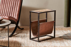The nkuku Alera Side Table pairs an iron frame and wood top with a brown leather magazine sling, styled on a woven rug next to a tufted brown leather chair and round side table.