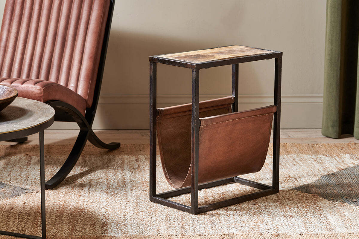 The nkuku Alera Side Table pairs an iron frame and wood top with a brown leather magazine sling, styled on a woven rug next to a tufted brown leather chair and round side table.