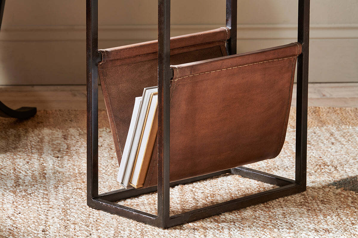 The Alera Side Table by nkuku features a brown leather magazine rack with a black iron frame, holding books and magazines. It sits on a woven beige rug in a room with light walls and wooden floors.