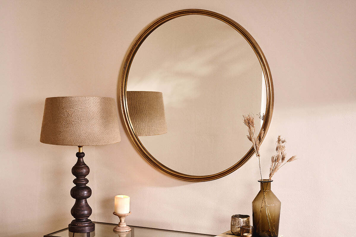 The Almora Round Mirror - Large by nkuku hangs on a cream wall above a glass table topped with a dark wooden lamp, beige shade, lit candle, green vase with dried grass, and small glass container. Soft light adds a warm, cozy ambiance.