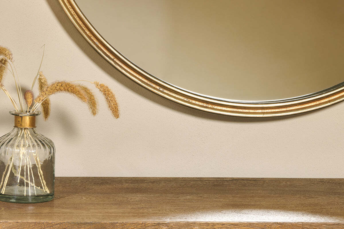 A close-up of a wooden table with a ribbed glass vase holding dried grasses. Above, the nkuku Almora Round Mirror - Large partially reflects light against a beige wall for a warm, minimalist look.