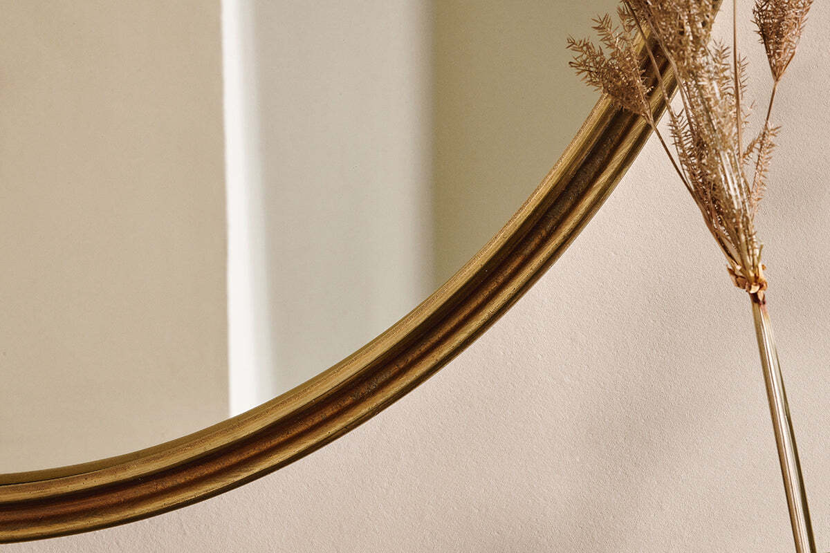 The nkuku Almora Round Mirror - Large, with its gold-toned frame, is positioned to the left, reflecting a light wall. To the right stands a spray of dried ornamental grass with golden-brown feathery plumes.