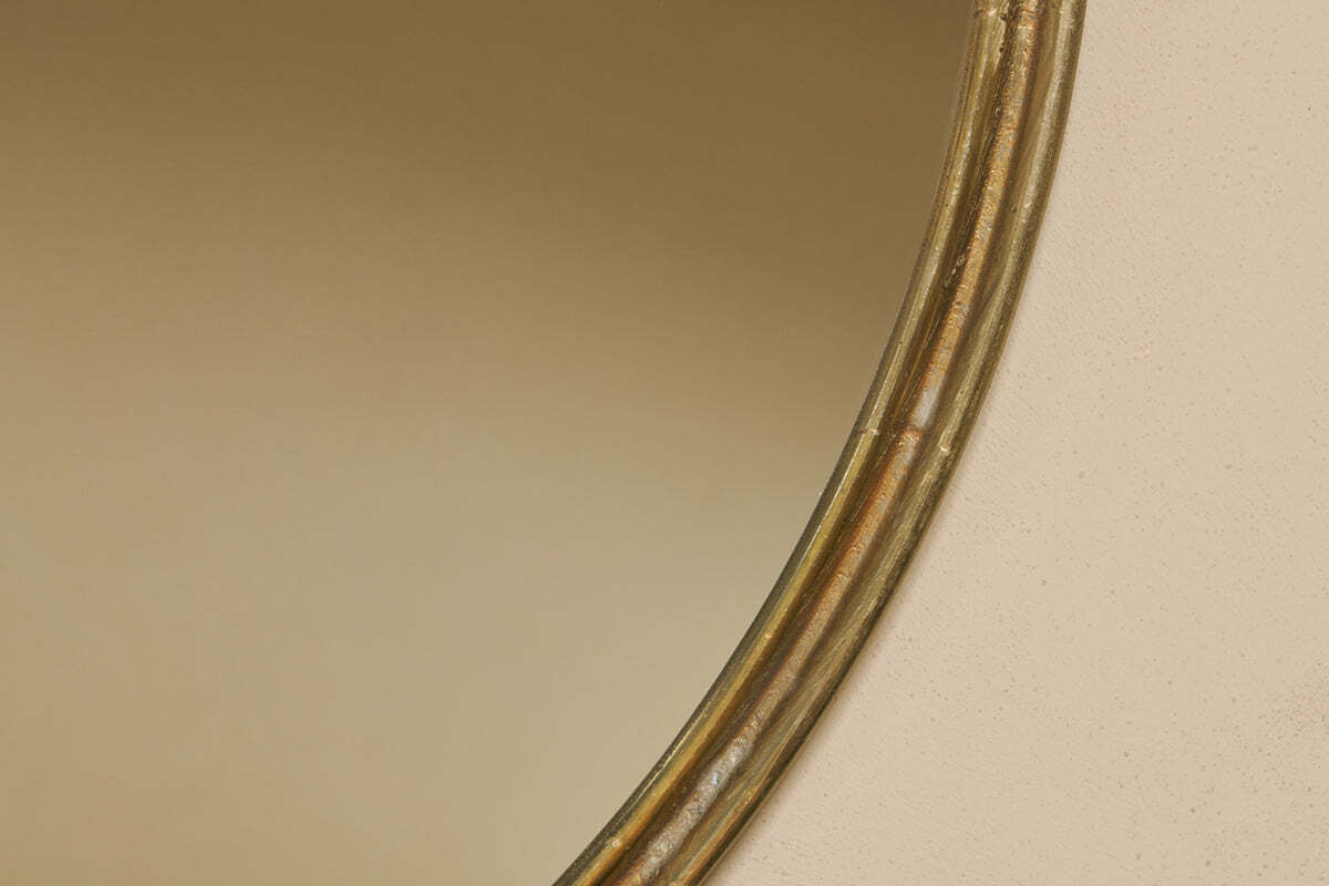 Close-up of the Almora Round Mirror - Large by nkuku, showing its gold-rimmed edge against a beige wall. The mirror’s curved frame and its soft reflection blend with the background, with only part of the frame visible on the right.