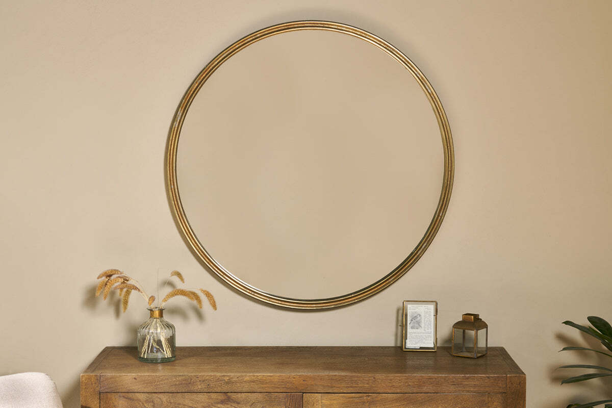 The Almora Round Mirror - Large by nkuku hangs above a wooden console table topped with a glass vase of dried grasses, a small framed picture, and a square lantern, creating a cozy, minimalist vibe against the neutral wall.