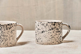 A set of two nkuku Ama Short Splatter Mugs with handmade black splatter designs sit on a light textured surface. Only part of the left mug is seen, the right mug is fully visible, both handles facing right against a neutral background.