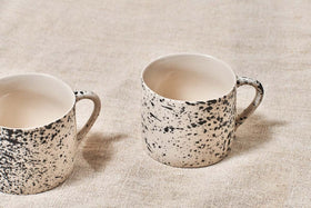 Two nkuku Ama Short Splatter Mugs (Set of 2) with black splatter patterns and white interiors sit on beige textured fabric. The mug on the right is fully shown, while the left is partly out of frame; both include handles.