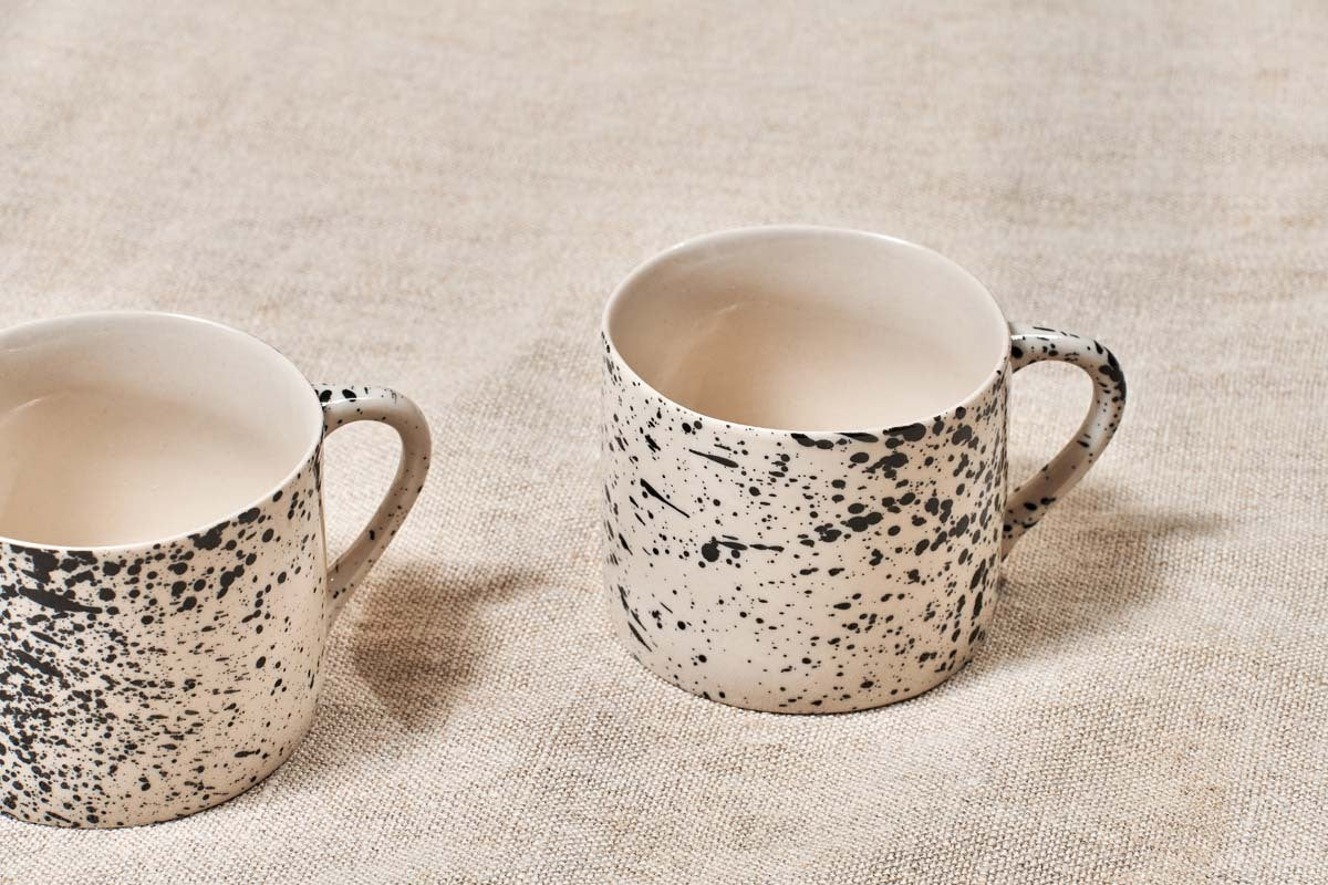 Two nkuku Ama Short Splatter Mugs (Set of 2) with black splatter patterns and white interiors sit on beige textured fabric. The mug on the right is fully shown, while the left is partly out of frame; both include handles.