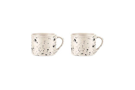 The Ama Short Splatter Mug Set by nkuku features two white handmade mugs with black splatters, rounded bodies, and large handles, displayed side by side on a plain white background in a minimalist, well-lit photo.