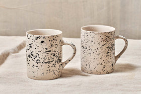 Two nkuku Ama Mug - Tall ceramics with a black splatter finish stand side by side on textured, light fabric. The softly blurred, neutral background highlights the artistic, handcrafted design of these unique mugs.