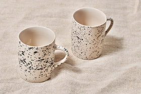 Two cream-colored nkuku Ama Mug - Tall mugs with bold black splatter finish rest on textured beige fabric. Both handmade mugs feature handles and a similar design, with the left one placed slightly closer to the camera than the right.