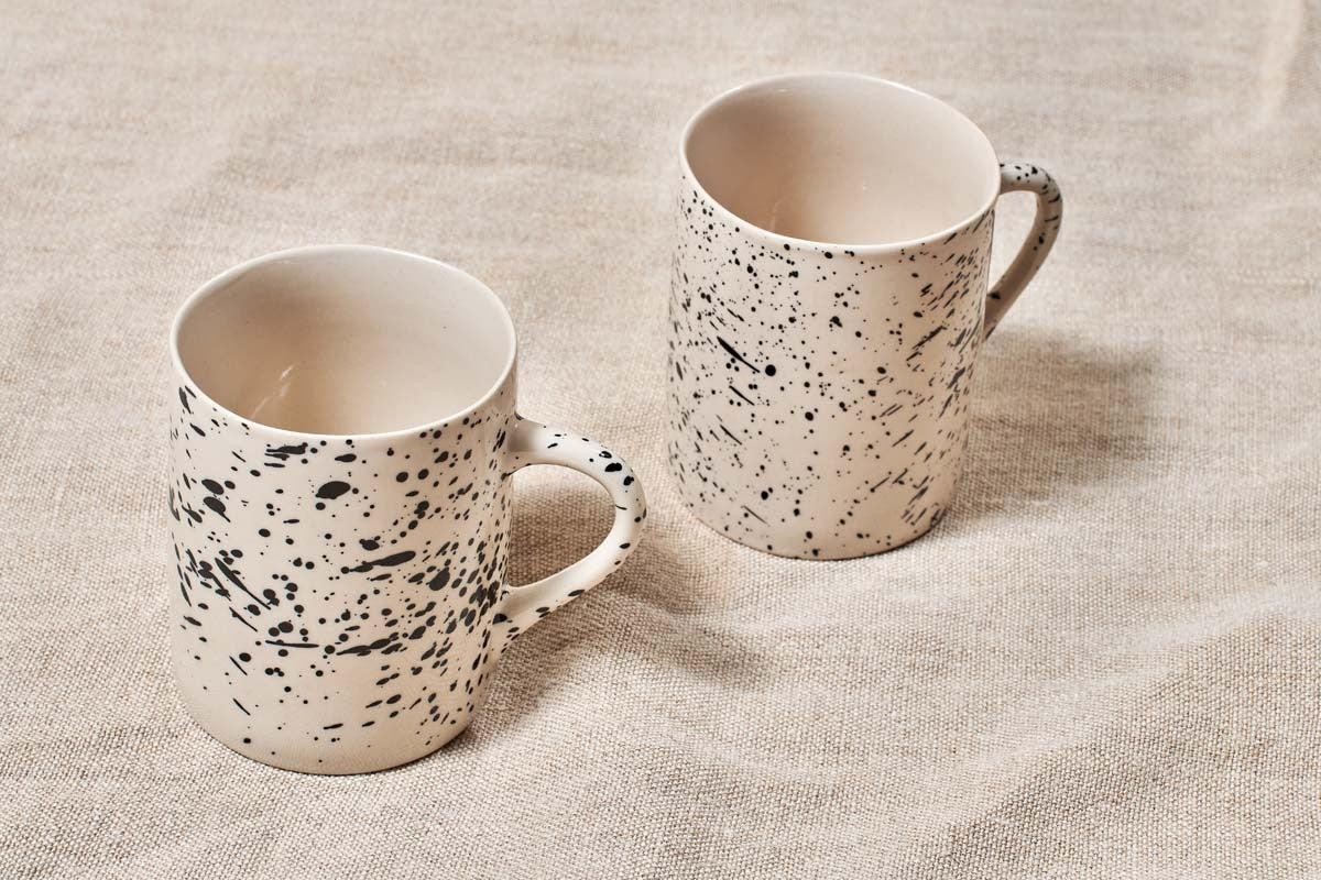 Two cream-colored nkuku Ama Mug - Tall mugs with bold black splatter finish rest on textured beige fabric. Both handmade mugs feature handles and a similar design, with the left one placed slightly closer to the camera than the right.