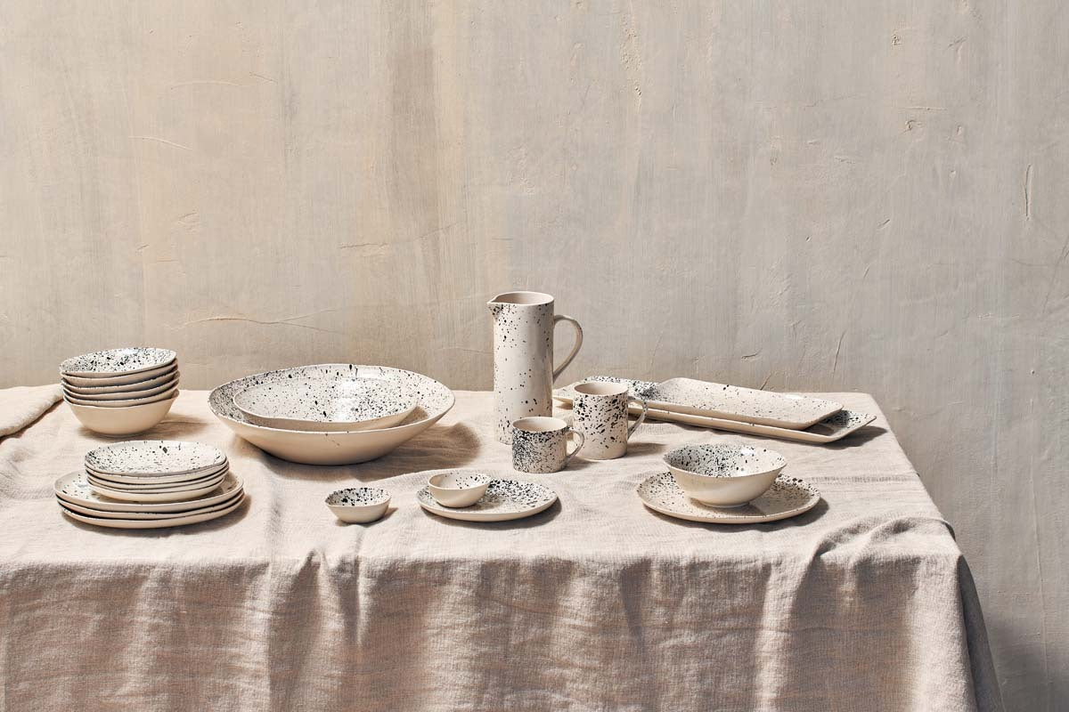 The nkuku Ama Mug - Tall features a cream-colored ceramic with a black splatter finish, perfect alongside matching bowls, plates, and a pitcher on a textured linen tablecloth.