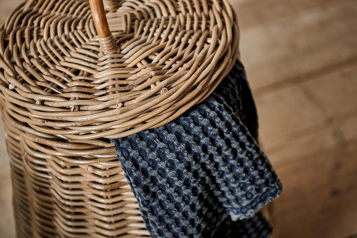 A close-up of the nkuku Amandi Rattan Laundry Bin with a curved handle. A textured, dark gray honeycomb towel is draped over its edge—an elegant storage solution. Wooden flooring appears softly blurred in the background.