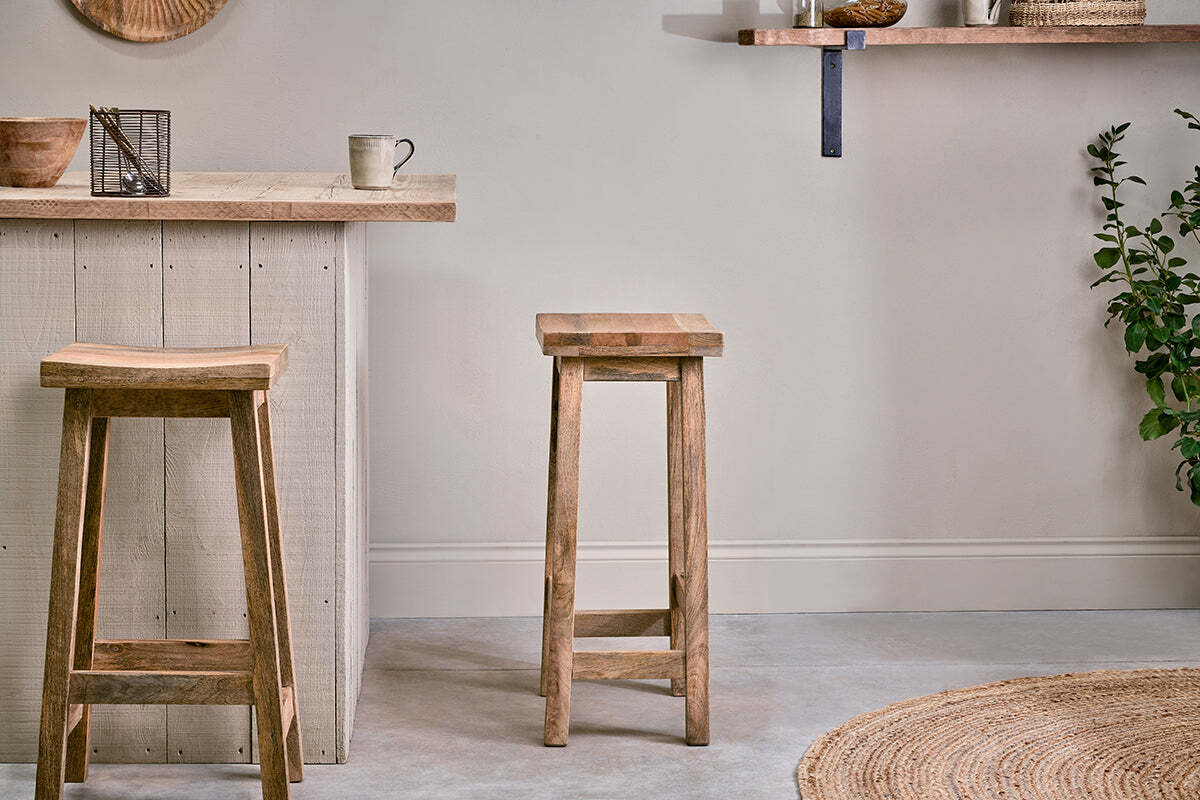Two nkuku Ambi Wooden Stool - Tall stools sit by a rustic light wood island. A beige mug and decor rest on top. A round woven rug is spread over the smooth floor, while a leafy plant stands to the right against a pale wall.