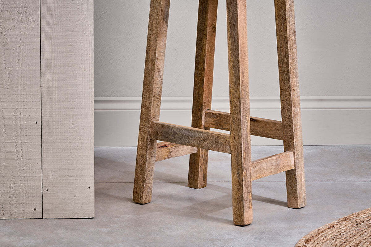 A close-up of the Ambi Wooden Stool - Tall by nkuku shows its handcrafted mango wood legs and natural grain on a smooth gray floor, with part of a textured cream table and woven rug nearby against a plain wall with molding.