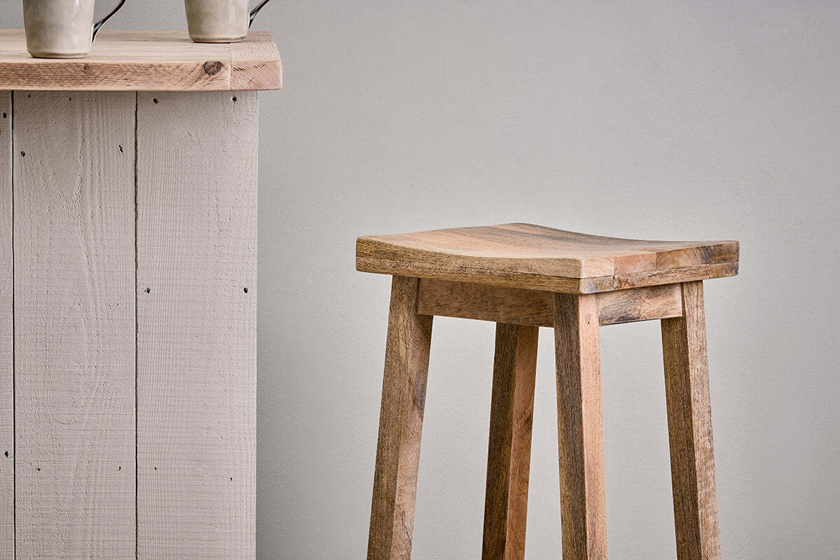 The nkuku Ambi Wooden Stool - Tall, featuring a curved mango wood seat and sturdy legs, stands beside a rustic light wood counter. Two handled ceramic mugs sit on the counter against a plain light gray wall.