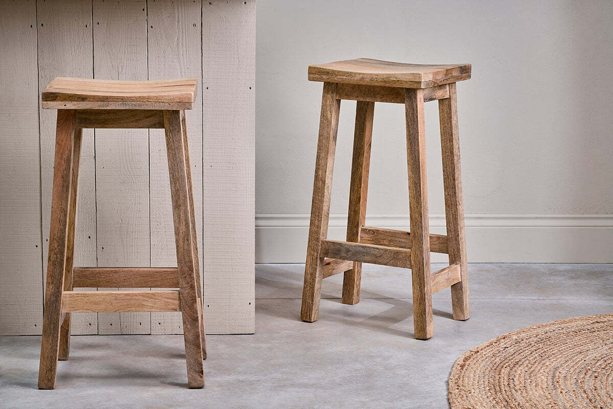 Two nkuku Ambi Wooden Stools - Tall, with curved mango wood seats, stand on a light-gray floor; one is by a light beige counter and the other close by. A round woven rug sits to the right against a pale gray wall.