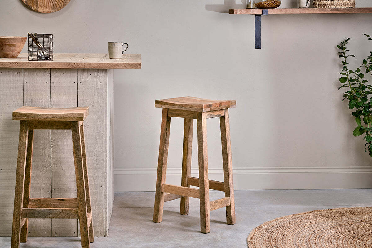 A minimalist kitchen featuring two nkuku Ambi Wooden Stools - Tall by a light wood counter. A mug, decor, woven rug, green plant, and a floating shelf with jars and bowls add warmth to the neutral space.