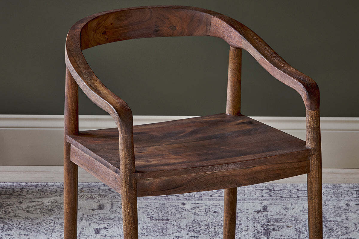 The Anbu Acacia Dining Chair by nkuku, in a washed walnut finish, features a curved backrest and armrests with smooth edges. It’s shown on a gray and white patterned rug against a dark green wall with a white baseboard.