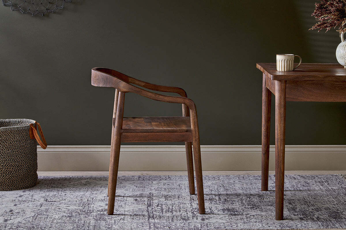 The nkuku Anbu Acacia Dining Chair in Washed Walnut sits on a patterned rug beside a dark wooden table holding a mug.