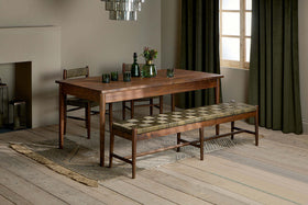 The Anbu Acacia Dining Table - Washed Walnut by nkuku sits on a rug, paired with matching chair and padded geometric bench. Green glassware, vases, emerald curtains, light wood flooring, and neutral walls complete the cozy look.