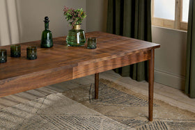 The nkuku Anbu Acacia Dining Table in Washed Walnut displays three green glass tumblers, a green bottle, and a clear vase with flowers on a beige patterned rug near green curtains and a sunlit window.