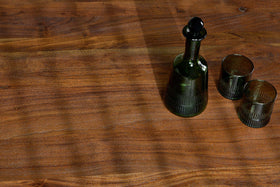The Anbu Acacia Dining Table in Washed Walnut by nkuku features a green glass decanter with a stopper and two matching tumblers. Diagonal shadows from window blinds fall across the wood, with the set arranged in the upper right of the image.